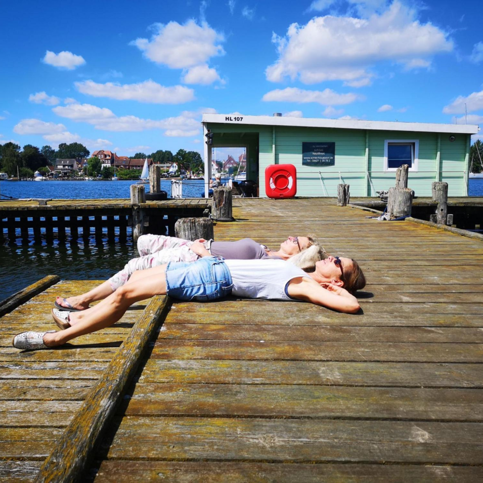 Das Bild zeigt zwei Menschen, die sich liegend auf einem Steg entspannen, im Hintergrund ein Hausboot und die Schlei.