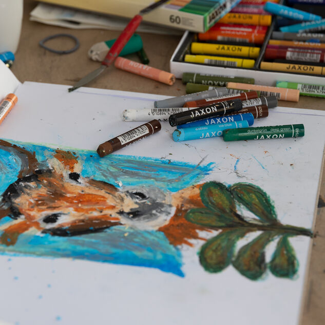 Das Bild zeigt farbige Kreiden und ein Blatt auf dem gemalt wird. Ein Tier mit Hörnern, gemalt mit Wachsmalstiften