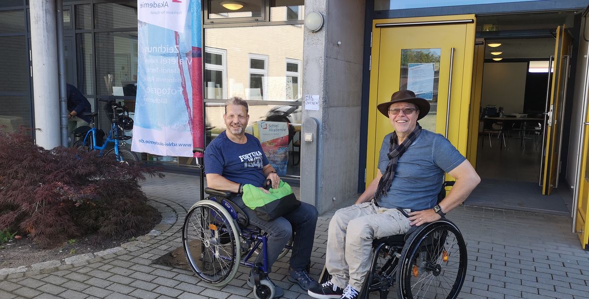 das Bild zeigt zwei Rollstuhlnutzer vor dem Haupteingang der Akademie.