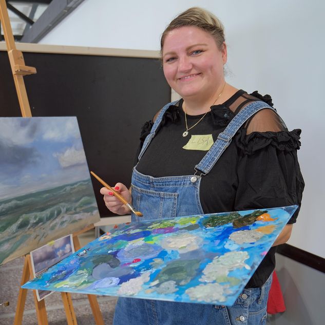 Eine Malerin hält eine Palette in der Hand und lacht in die Kamera