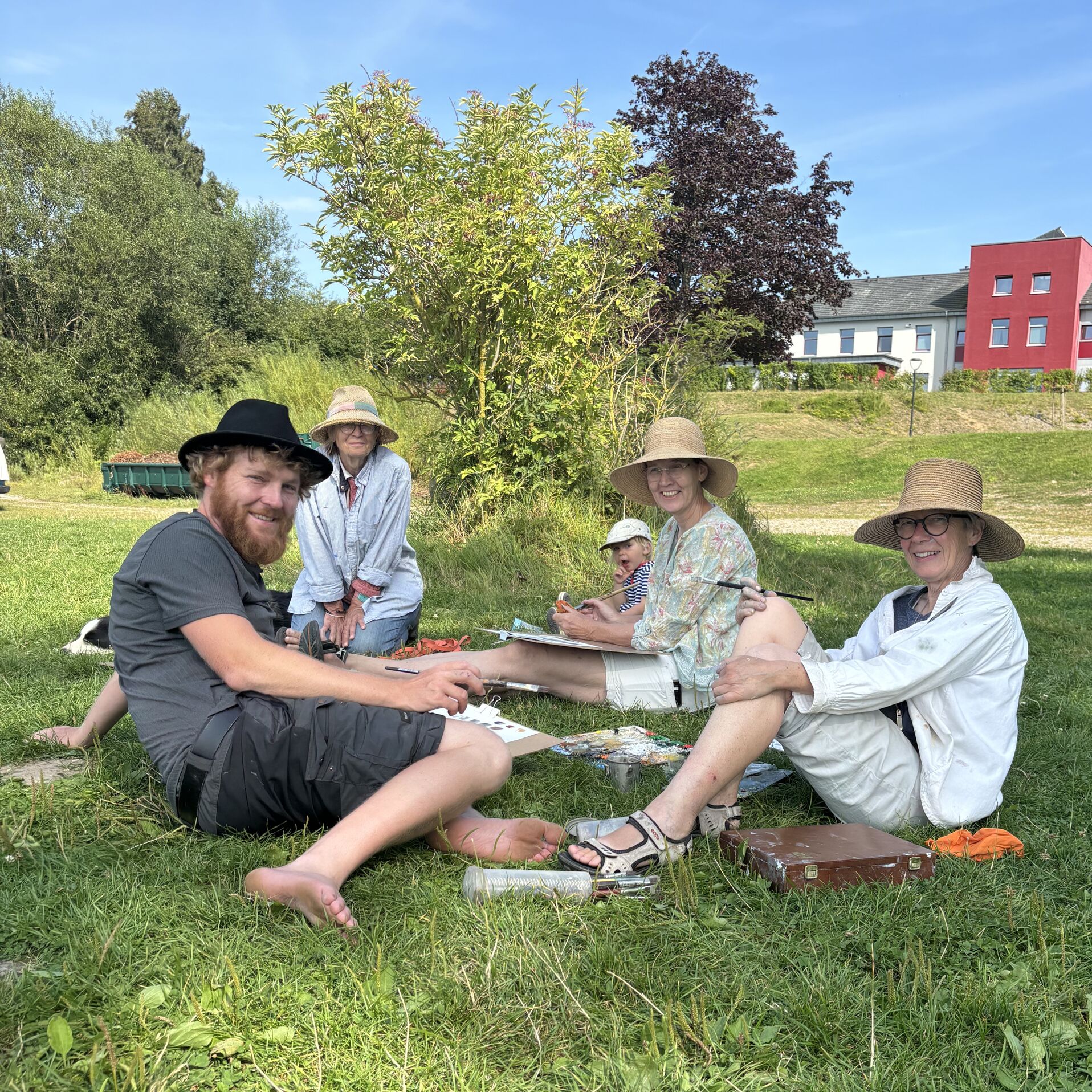 Eine Gruppe Menschen sitzt entspannt auf einer Wiese und lächelt in die Kamera. Die Menschen malen gerade Plein Air.