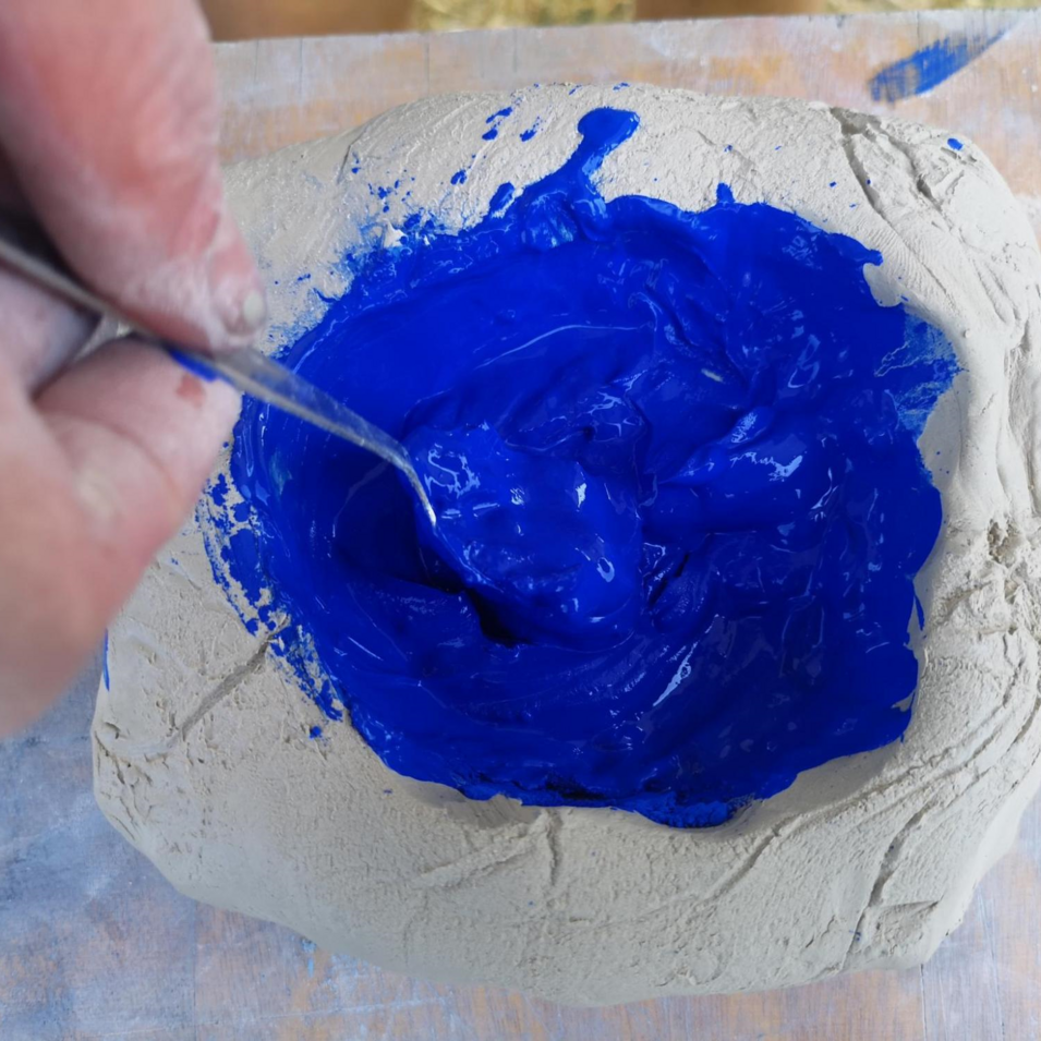 Das Foto zeigt eine Hand, die blaue Farbpigmente in den Ton mischt.